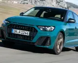 Audi RS1 verde azulado en movimiento por una carretera rural con montañas al fondo.