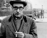 William Burroughs, con cappello e occhiali, tiene una sigaretta in mano, passeggiando in una strada urbana con persone sullo sfondo.