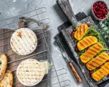 Dwa okrągłe sery camembert grillowane na ruszcie, obok pieczywo i grillowane kawałki sera na desce z granatem.