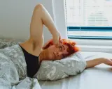 Jeune femme aux cheveux roux allongée dans son lit, le bras levé, le regard perdu. Elle semble fatiguée, peut-être un des symptômes manque magnésium.