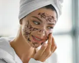 Une femme souriante applique un masque d'exfoliation visage. Elle porte une serviette sur la tête et un peignoir blanc.