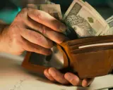 Dłonie wkładają banknoty 100 zł do brązowego portfela. To może być zapłata lub rekompensata.
