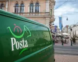 Zielona furgonetka pocztowa z logo "Posta" stoi na ulicy w węgierskim mieście. W tle widać kawiarnie i zabytkowe budynki.