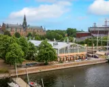Junibacken, ett museum tillägnat Astrid Lindgren, ligger vid vattnet i Stockholm, med en stor byggnad i bakgrunden och segelfartyg i hamnen.