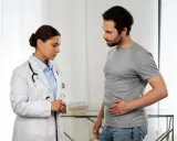 Lekarka w białym kitlu rozmawia z pacjentem, który trzyma się za brzuch. To może być początek rozmowy o tym, jak wygląda badanie urologiczne u mężczyzn.