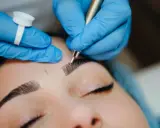 Main d'une esthéticienne en bleu, effectuant un entretien microblading sur les sourcils d'une cliente.