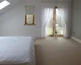 Dormitorio con cama blanca, ventana abuhardillada y puerta acristalada con cortinas. La orientación oeste de los pisos permite una luz suave.