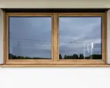 Nowoczesne okno z drewnianą ramą i parapetem zlicowanym ze ścianą. W odbiciu widać niebo i drzewa.