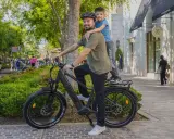 Ojciec z synem na rowerze elektrycznym, który wygląda jak jazda rowerem elektrycznym bez wspomagania. Słońce, zieleń i sklepy w tle.