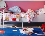 Habitación infantil con dos camas blancas, cojines de nube y mantas coloridas. Alfombra azul con tren de juguete y vías.