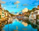 Un pintoresco pueblo pesquero con casas blancas y verdes, barcos amarrados en un canal de aguas cristalinas y un cielo azul con nubes esponjosas.
