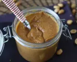Un pot de praliné cacahuète onctueux, prêt à être dégusté. Quelques cacahuètes parsèment le plan de travail.