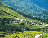 Un villaggio alpino nel **Pian della Regina**, con case in pietra e un fiume che scorre tra prati verdi e boschi lussureggianti.