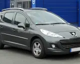 Szary Peugeot 207 1.4 benzyna z relingami dachowymi, zaparkowany przed niebieskim budynkiem.