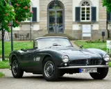 Un elegante BMW descapotable gris, un clásico de Alemania, aparcado frente a un edificio histórico con árboles en flor.