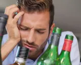 Mężczyzna z ręką na czole, otoczony butelkami, wyraźnie cierpi z powodu objawów przepicia alkoholowego.