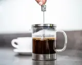 Eine Hand drückt den Stempel in eine French Press, um den perfekten Kaffee-Brew zuzubereiten. Eine Tasse wartet im Hintergrund.