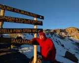 Mężczyzna w czerwonej kurtce przy znaku "Mount Kilimanjaro", najwyższy szczyt Afryki. Gratulacje, jesteś na Stella Point 5756 m n.p.m.