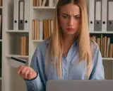Jeune femme perplexe devant son ordinateur, carte bancaire à la main, craignant une arnaque location appartement.