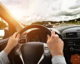 Ręce na kierownicy, czuć lekkie bicie kierownicy przy hamowaniu na drodze. Słońce przebija się przez chmury.