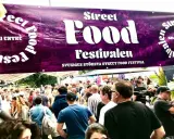 Stor folkmassa vid Sveriges största street food festival. En lila banderoll med texten "Street Food Festivalen" hänger över besökarna.