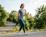 Frau genießt **incline walking** auf einem asphaltierten Weg, umgeben von grünen Bäumen. Sie hält eine Wasserflasche in der Hand.