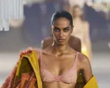 Modella in passerella con un audace reggiseno a vista color pesca, abbinato a una giacca gialla con pelliccia arancione.