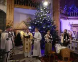 Pasterka w kościele w Zakopanem. Ksiądz czyta z księgi przy choince, obok ministranci i wierni w tradycyjnych strojach.