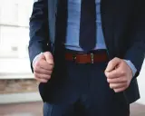 A man in a navy pinstripe suit and blue shirt wears a brown belt with black pants.