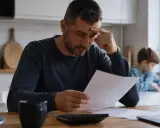 Zmartwiony mężczyzna przegląda dokumenty, licząc koszty utrzymania dziecka. W tle widać syna.
