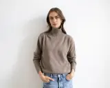 Kobieta w beżowym golfie z wełny merino i jeansach, z rękami w kieszeniach, na białym tle.