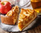 Deux parts de tarte aux pommes sur une ardoise, saupoudrées de sucre glace, avec des pommes et un verre de cidre.