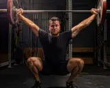 Ein Mann im tiefen Squat mit einer Langhantel über dem Kopf. Seine Muskeln sind angespannt, bereit für den nächsten Schritt im **power lifting**.