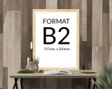 Ramka z napisem "FORMAT B2 707mm. x 500mm." na tle drewnianej ściany. Na stole obok świece, rośliny i otwarta książka.