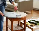Une personne en jean et chemise à carreaux, portant des gants, utilise un rouleau pour appliquer une peinture grise sur une table ronde en bois. Le **décapage bois** est en cours pour un relooking.