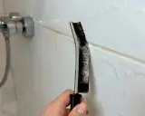 Hand reinigt Duschfugen mit einer Bürste und Schaum.