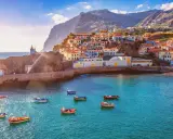 Bunte Häuser schmiegen sich an steile Hänge auf den Inseln bei Madeira. Kleine Boote schaukeln im azurblauen Wasser vor einem Leuchtturm.