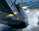 Un skipper manœuvre sur l'IMOCA Bureau Vallée, le bateau noir et jaune fend les vagues dans un embrun spectaculaire.
