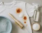 Cómo quitar mancha de óxido en la ropa: una camiseta blanca con manchas marrones, un cepillo, un bol con agua jabonosa, un spray y bicarbonato.