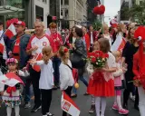 Dzieci i dorośli w strojach ludowych i z polskimi flagami maszerują ulicą. Widać serca z czerwonych balonów i kwiatów. Polacy w Szwecji świętują.
