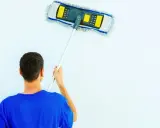 Hombre con camiseta azul limpiando paredes blancas con un plumero.