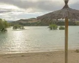 Sombrilla de paja en la orilla del pantano de Alloz. El agua turquesa, árboles emergiendo y colinas al fondo invitan a descubrir cómo llegar.