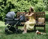 Kobieta w żółtej sukience siedzi na ławce obok wózka Kinderkraft. Piesek leży na trawie.