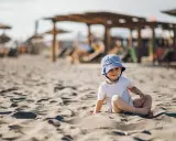 Uroczy maluch w pasiastym kapelusiku siedzi na piaszczystej plaży. Idealne miejsce, gdzie na wakacje z rocznym dzieckiem.