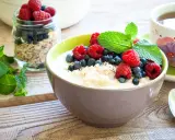 Schüssel mit Haferflocken mit Kefir, garniert mit Himbeeren, Blaubeeren und Minze. Daneben ein Glas Müsli und eine Tasse Tee. Gesundes Frühstück.