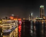 Beleuchtete Nilschiffe und moderne Hochhäuser spiegeln sich im Wasser. Ein magischer Anblick von Kairo bei Nacht.