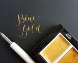 Złoty napis "Blue Gold" obok pióra i palety farb.