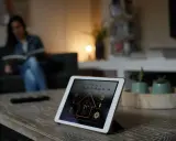 Tablette affichant l'interface pour installer un thermostat connecté. Une femme lit sur le canapé en arrière-plan.