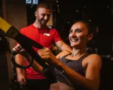 Frau lacht und trainiert mit TRX-Bändern, ihr Trainer lächelt. Sie will schnell wieder fit werden.