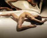 Młoda kobieta w cielistym body wygina się w łuk na podłodze, jej odbicie w lustrze tworzy surrealistyczny obraz. Jak z opowiadanie erotyczne nastolatki.